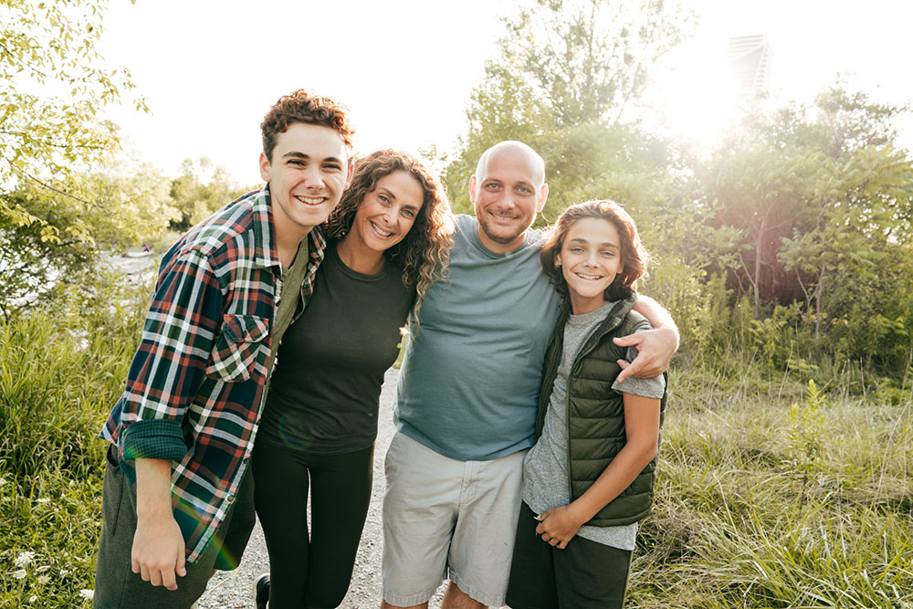 A white family (a teenager, a tween, and their mom and dad) smile at the camera outdoors. They are in light activewear and are hiking.