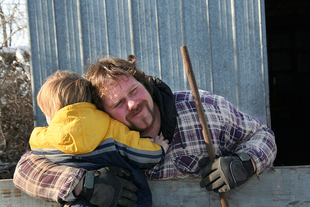 A white farmer holding the handle of a garden implement hugs his toddler son with his other arm, leaning over a wooden fence to reach him. The toddler wears a bright yellow striped windbreaker and his father wears a heavyweight flannel shirt with a hood and sturdy gloves.