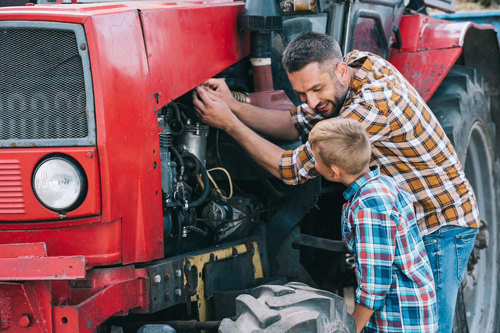 A white father and son work together on the engine of a tractor. The son observes as his father explains. Both are wearing colorful plaid shirts.