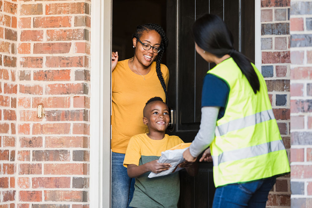 A Black mother and her elementary-aged son greet a postal worker at the door of a brick house. The son smiles as he accepts the mail.
