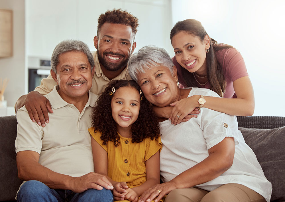 A multigenerational mixed-race family (an elementary-aged child, her parents, and her grandparents) pose together on the couch and smile for the camera.