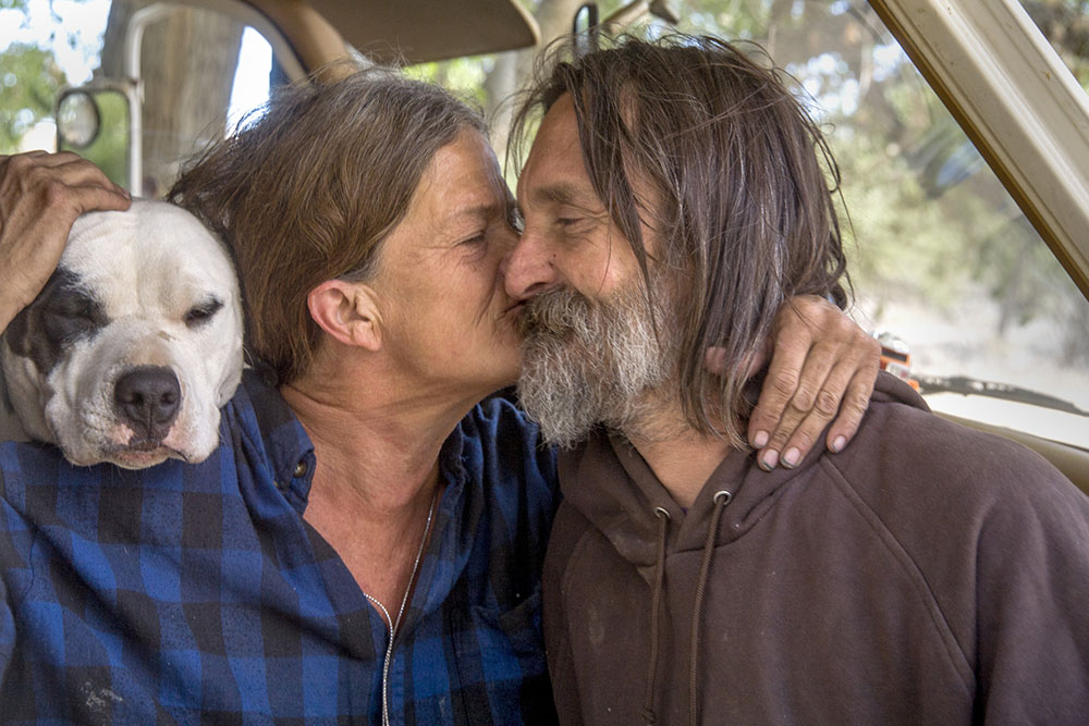 An affectionate indigent couple and their dog live in a truck camper among homeless residents of a primitive outdoor encampment in the desert town of Victorville, CA.