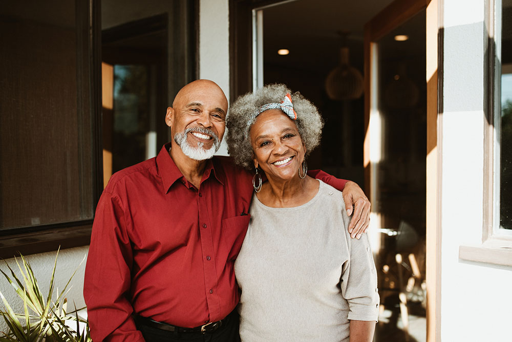 A retired Black couple on their front porch. The husband wears a red button-down shirt. The wife wears an oatmeal-colored light sweater and a coordinating plaid headband with a bow.