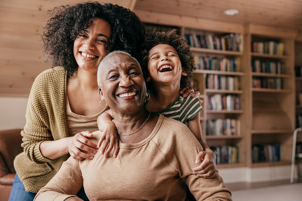 Three generations of Black women (a child, a mother, and a grandmother) wear coordinating colors and smile at the camera in a wood-paneled room with bookshelves behind them.