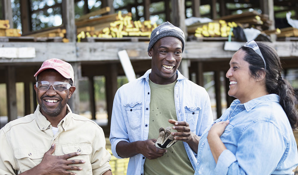 A multiracial group of three workers at a lumberyard, standing together, laughing. The Hispanic woman and the African-American man wearing the trucker's hat and safety glasses are in their 40s. The main focus is on the young African-American man standing in the middle. He is in his 20s. There are shelves stacked with lumber out of focus behind them.